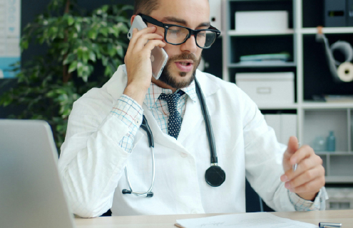 arzt-im-sprechstundenraum-telefoniert-und-arbeitet-am-laptop-im-medizinischen-umfeld